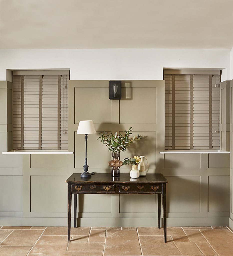 Venetian blinds closed in a hall space with a dark wood table framing the interior space. Natural colour scheme with wall panels and tiled flooring.