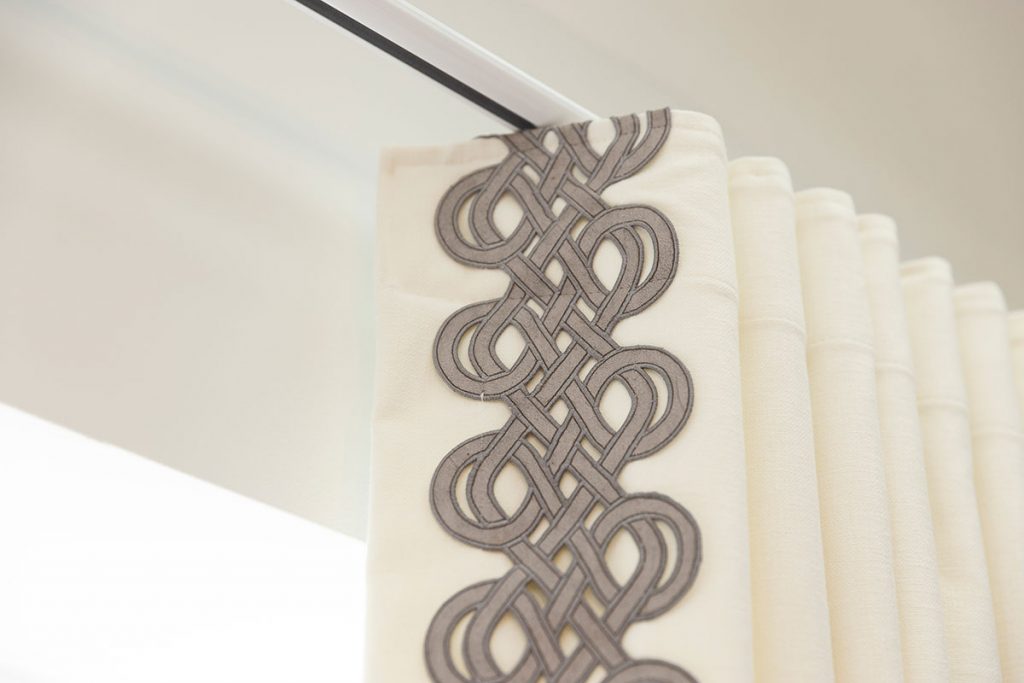 A close up shot of curtains edging with an embroidery pattern that is contemporary. Black and grey thread contrasts the white curtains. Shot exhibits the discrete track and ability to fit to ceiling spaces.